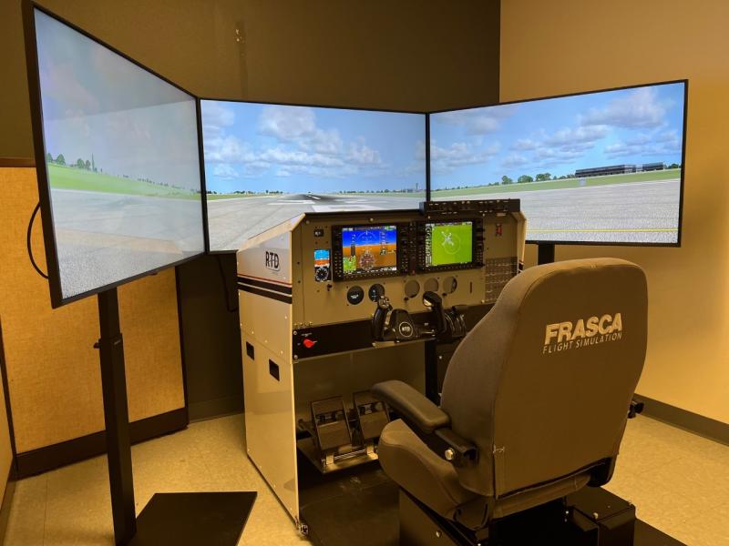 cwu-simulator consisting of chair, airplane control planels and switches in front of the thair; three monitors showing a simulated runway and blue, partially cloudy sky