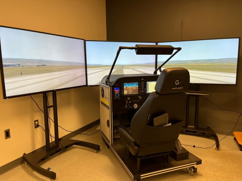 cwu-simulator consisting of chair, airplane control planels and switches in front of the thair; three monitors showing a simulated runway and blue, partially cloudy sky