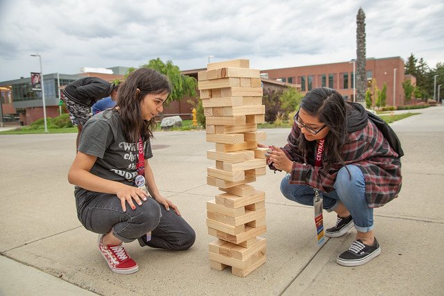 Two students playing Jenga while crouching on the concrete. CWU's Samuelson building is in the background, it's a cloudy day.