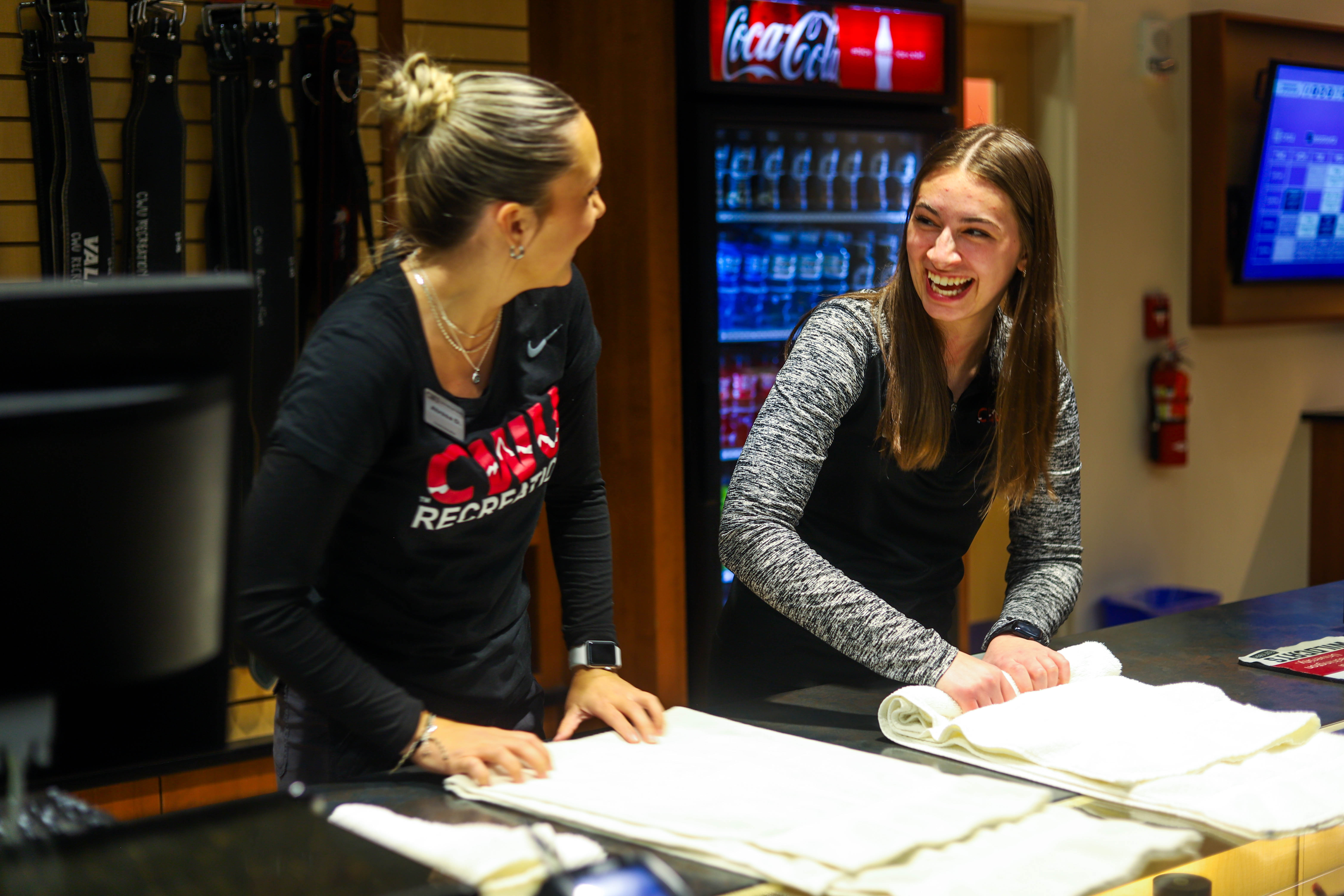 Two female student workers at the Recreation Center front desk laughing together and cleaning the counter. They are both wearing CWU gear.