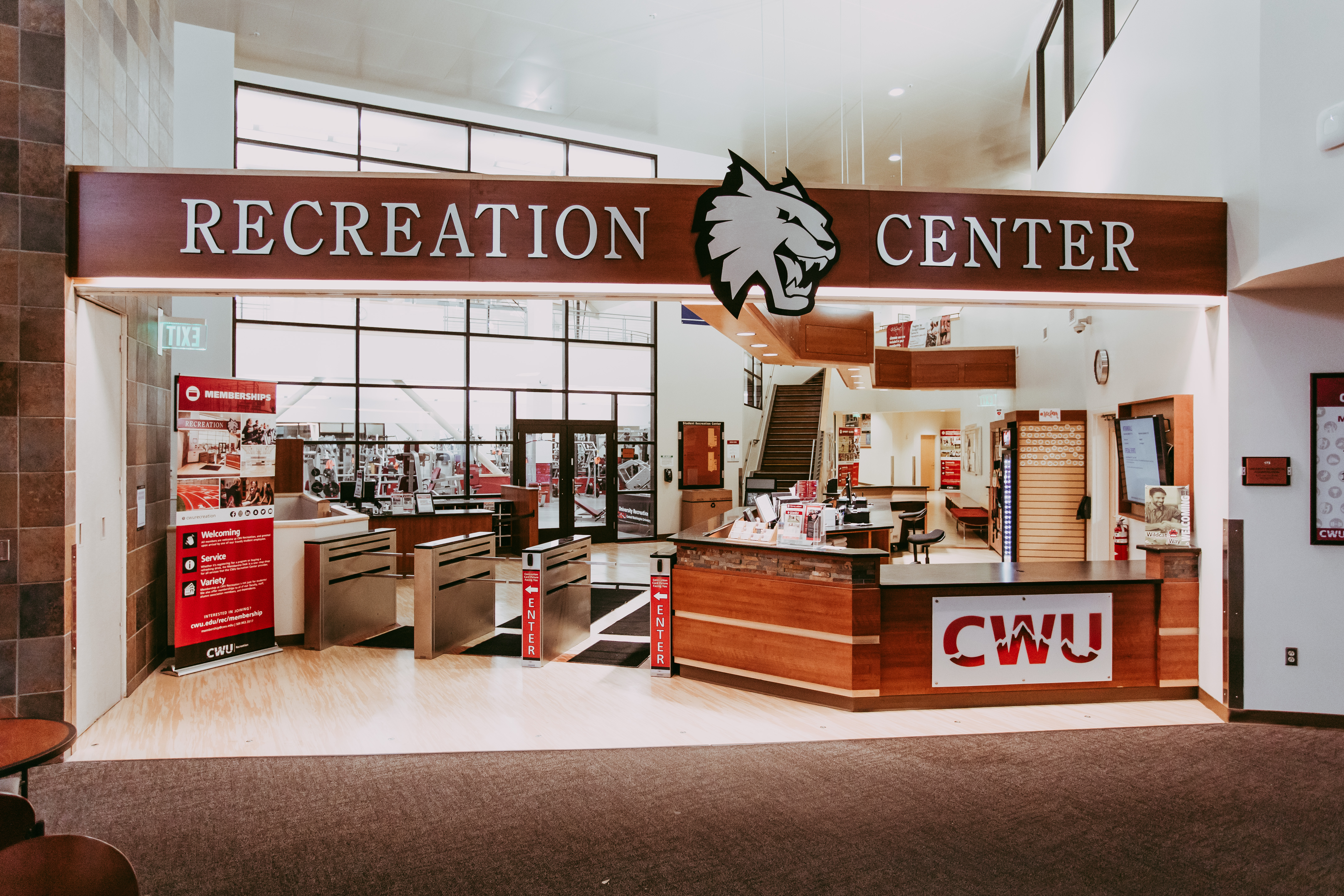 An image of the recreation center at CWU. There is a front desk and terminal to swipe your student card before entering. In the back there is a weight room and a staircase. Above the front desk there is a big sign that has the Wildcat logo and the words "RECREATION CENTER".